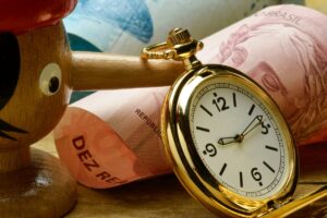 Relógio de bolso dourado apoiado sobre cédulas de reais enroladas, com destaque para as inscrições "República" e "Dez Reais", simbolizando a relação entre tempo e dinheiro no contexto financeiro brasileiro.